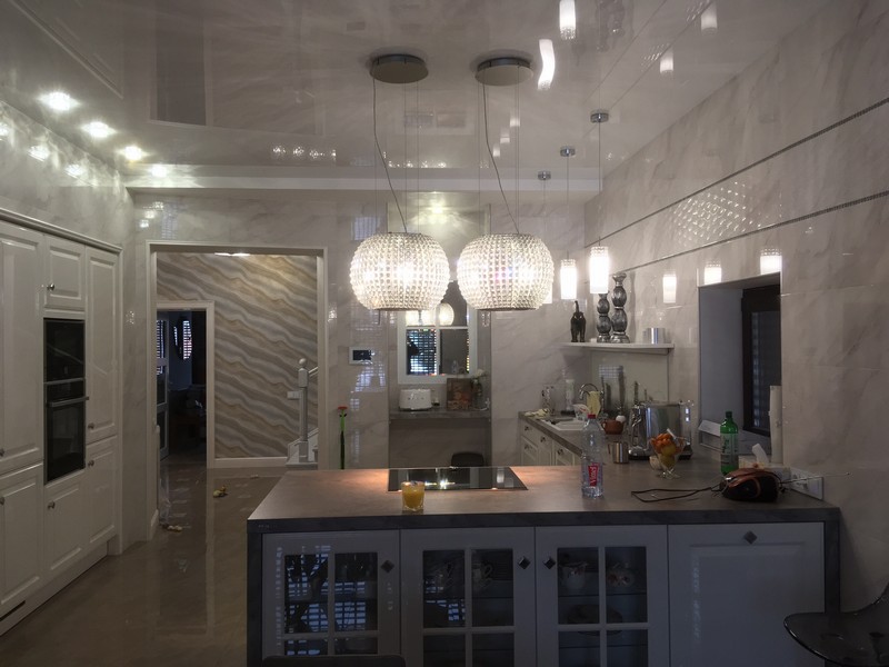 Белый лаковый натяжной потолок на кухне с двумя люстрами и светильниками по периметру.